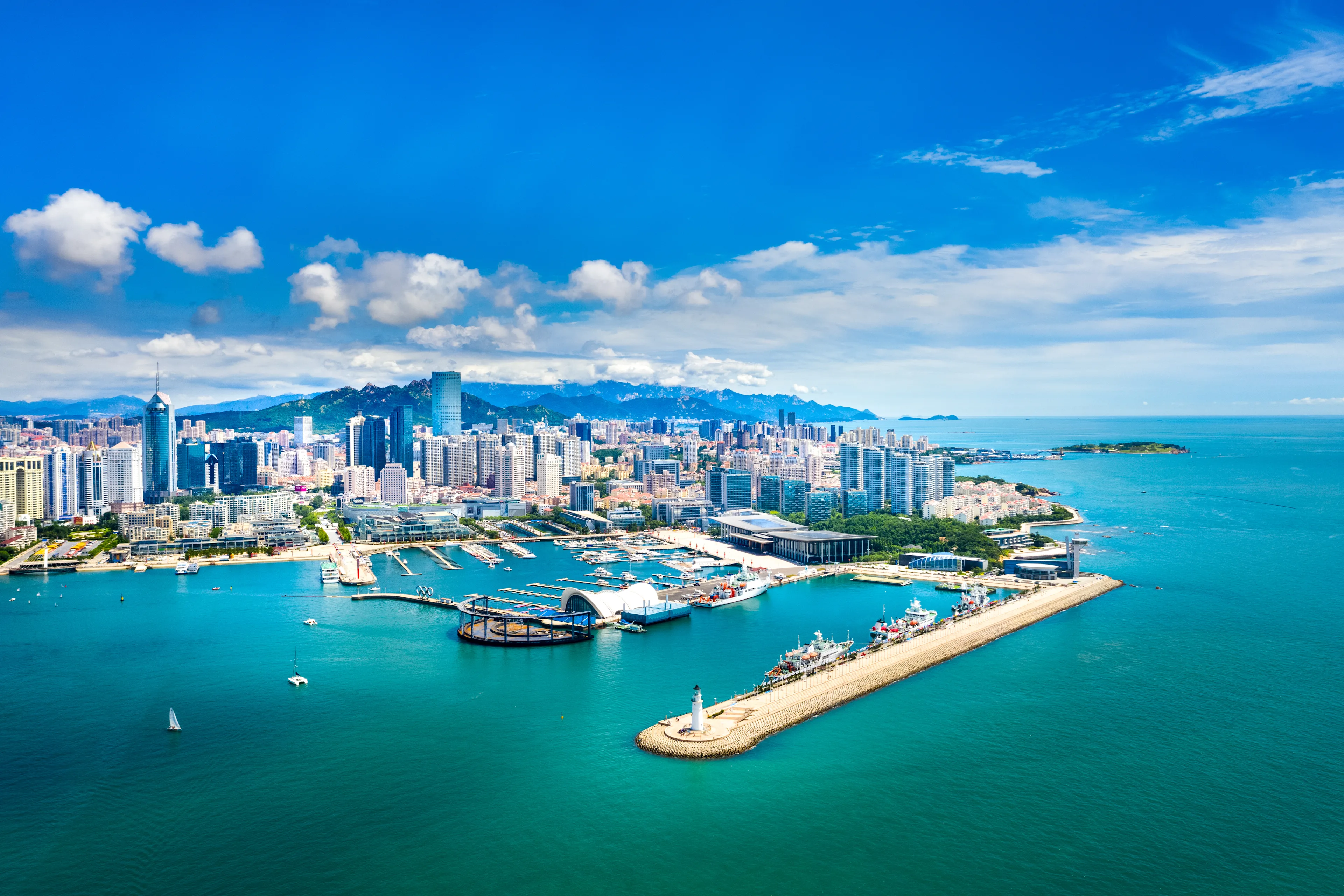 Aerial view of Qingdao, China, highlighting its modern skyline along the coastline. The Olympic Sailing Center is prominently featured by the waterfront, with its marina and sailing boats in the clear blue waters.