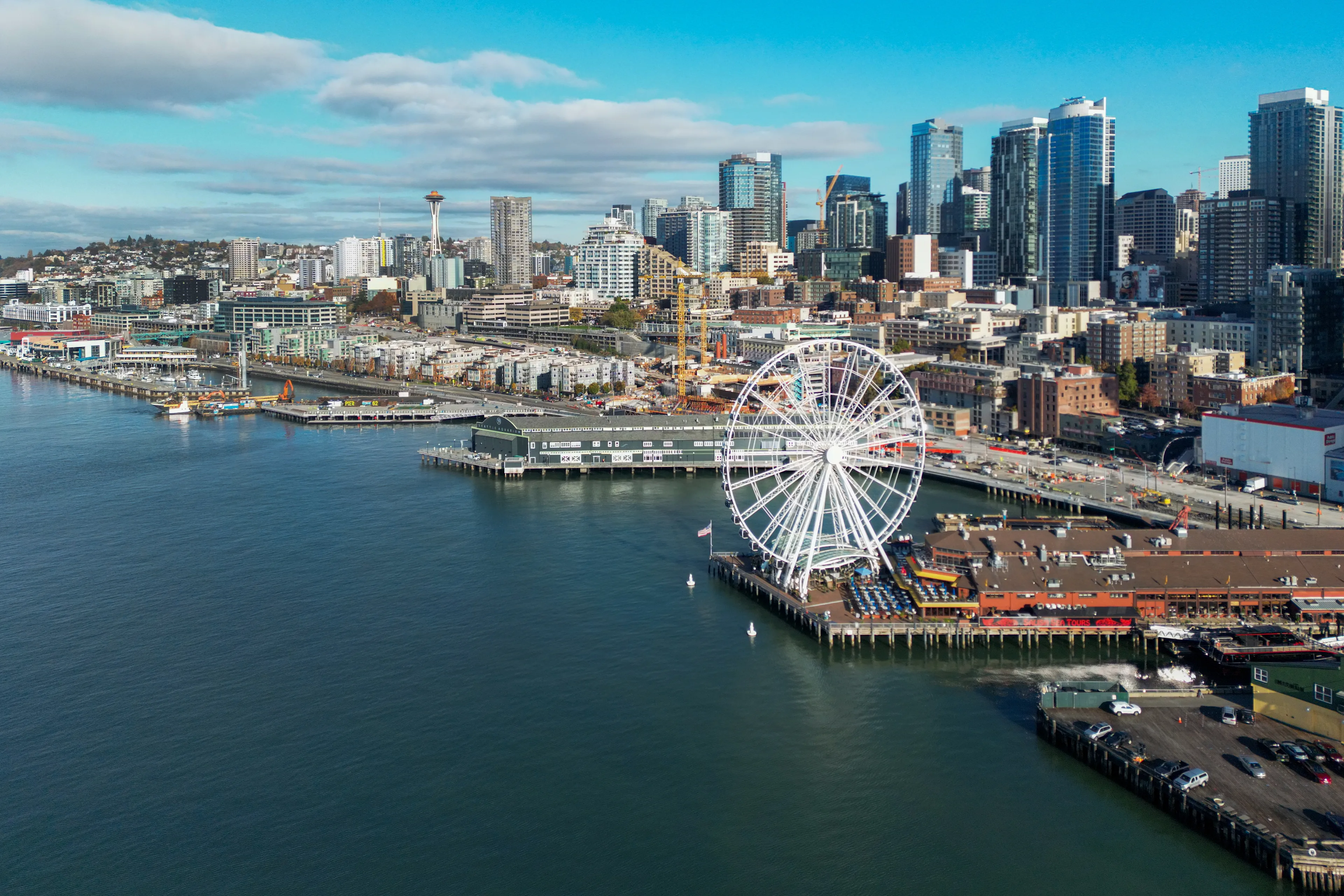 Aerial view of Seattle's Bell Harbor Marina, highlighting the Seattle Great Wheel and the city's skyline, including the iconic Space Needle along the bustling waterfront.